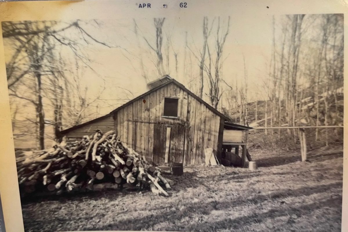 A sugarhouse beside a stacked woodpile, in a photograph dated April 1962.