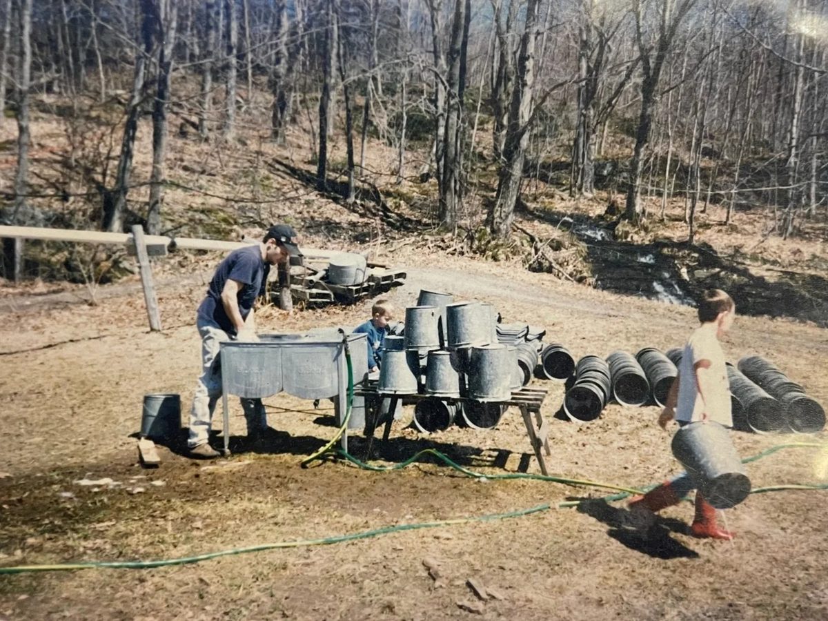 An adult and two children washing sap buckets outdoors at the edge of the woods.