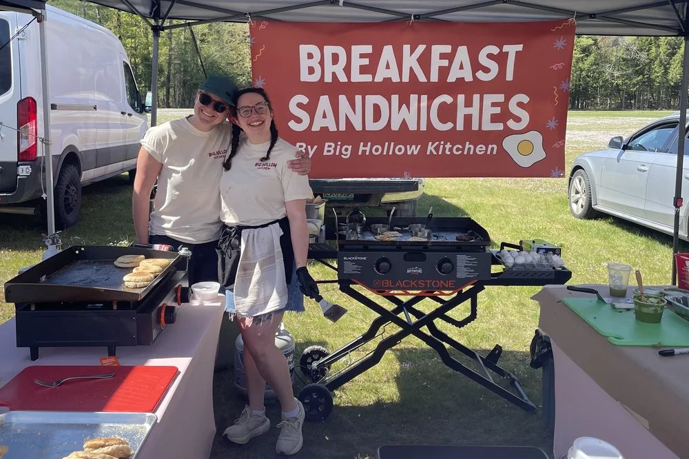 Kelly and Amanda Danckert side by side under their orange Breakfast Sandwiches banner, griddles either side of them at the market.