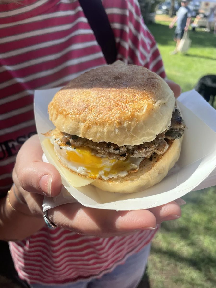 A breakfast sandwich held in a paper boat at the market — sausage, melted cheese and a fried egg with a soft yolk on a scratch-made English muffin.