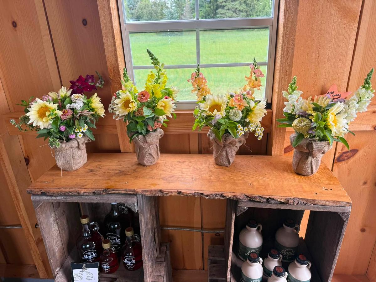 Wrapped bouquets lined up on a live-edge wooden bench inside the stand, with bottles of maple syrup and jars of honey on the shelf below.