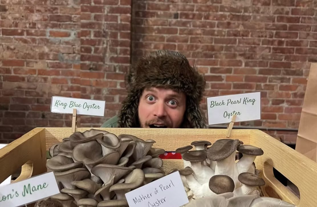 Blake Goldberg looking over a wooden crate of mushrooms with handwritten variety labels: King Blue Oyster, Black Pearl King Oyster, Mother of Pearl Oyster and Lion's Mane.